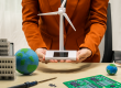 Business woman holding a model wind turbine with a repaired circuit board/PCB on the desk next to soldering iron and some integrated circuit chips. A metaphor for how electronics repair benefits renewable energy sustainability.