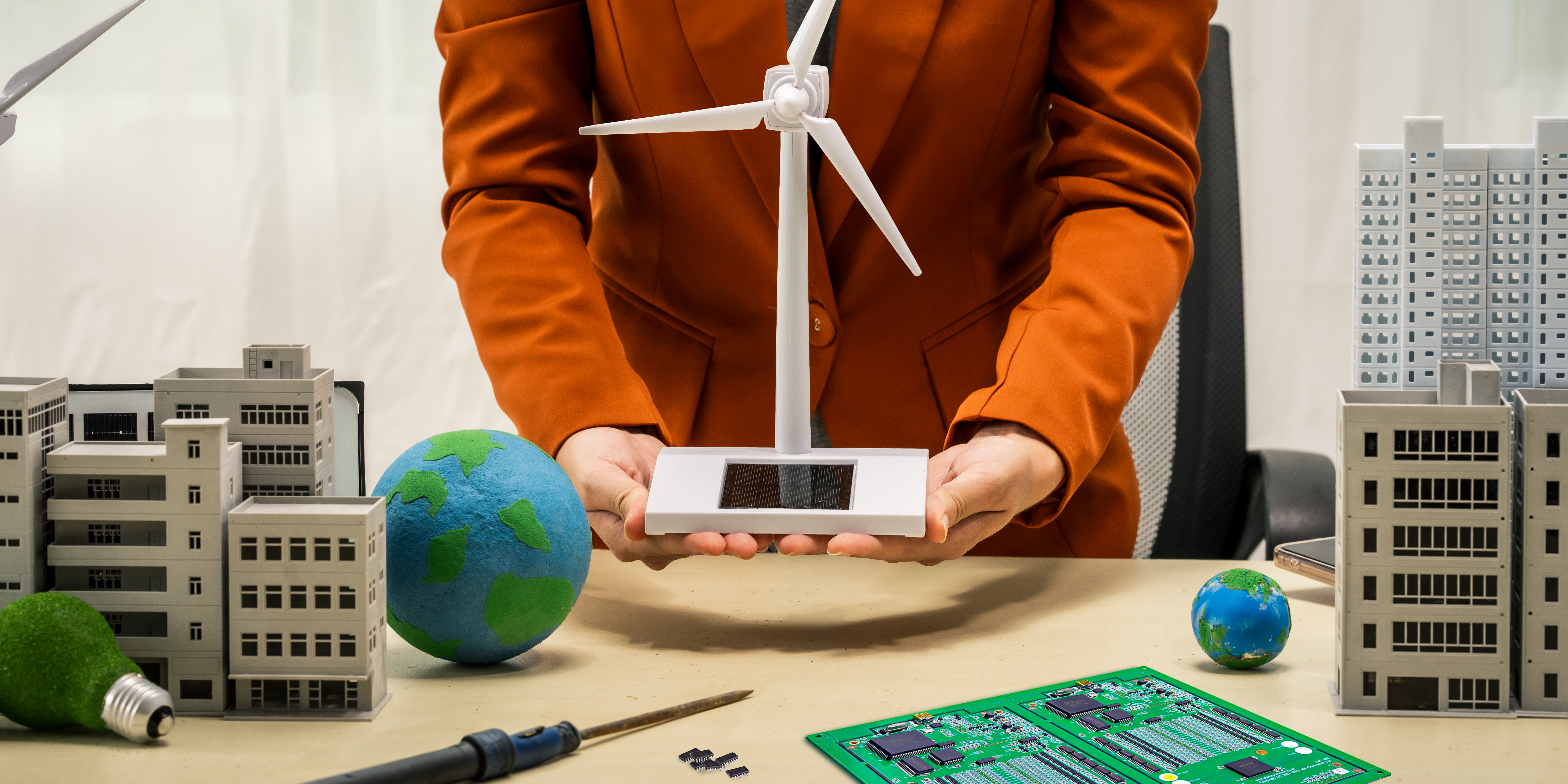 Repair in Renewable Energy Business woman holding a model wind turbine with a repaired circuit board/PCB on the desk next to soldering iron and some integrated circuit chips. A metaphor for how electronics repair benefits renewable energy sustainability.