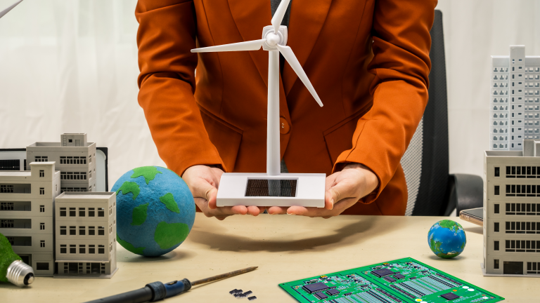 Business woman holding a model wind turbine with a repaired circuit board/PCB on the desk next to soldering iron and some integrated circuit chips. A metaphor for how electronics repair benefits renewable energy sustainability.