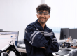 Smiling technician in safety glasses and navy overalls, arms crossed, at an industrial electronics repair bench with a computer and robotic arm