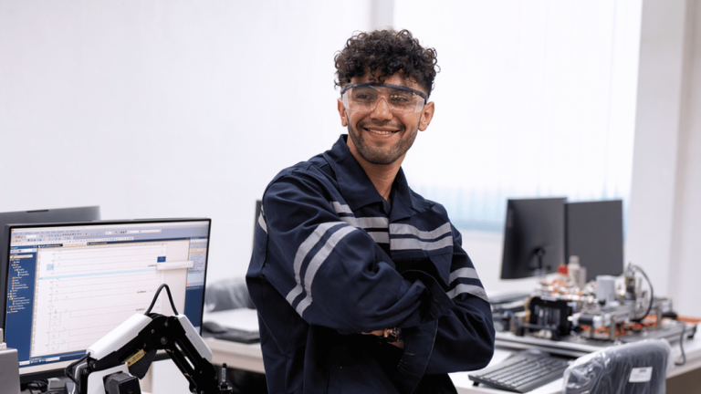 Smiling technician in safety glasses and navy overalls, arms crossed, at an industrial electronics repair bench with a computer and robotic arm